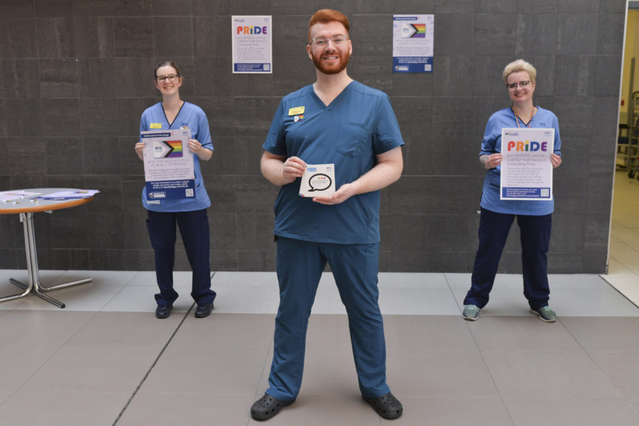 Amanda Law, Andrew Laird, and Katie Sharp of the NHS Greater Glasgow and Clyde LGBTQ+ Staff Forum. They are standing in the atrium at thr QEUH, holding posters.