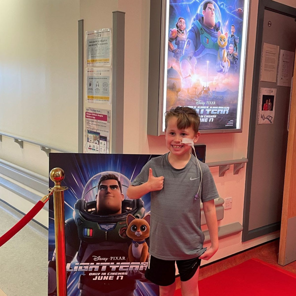 A young boy giving a thumbs up, standing next to an advert for Lightyear.