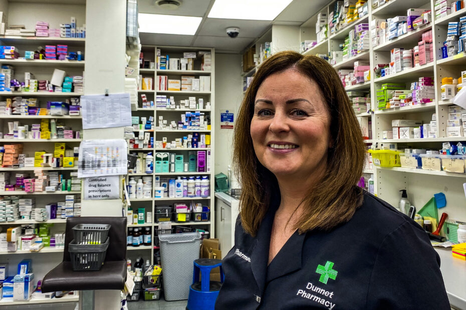 Maryann Dunnet of Dunnet Pharmacy in Glasgow, in the dispensing area of the pharmacy, with shelves of medicines behind her.