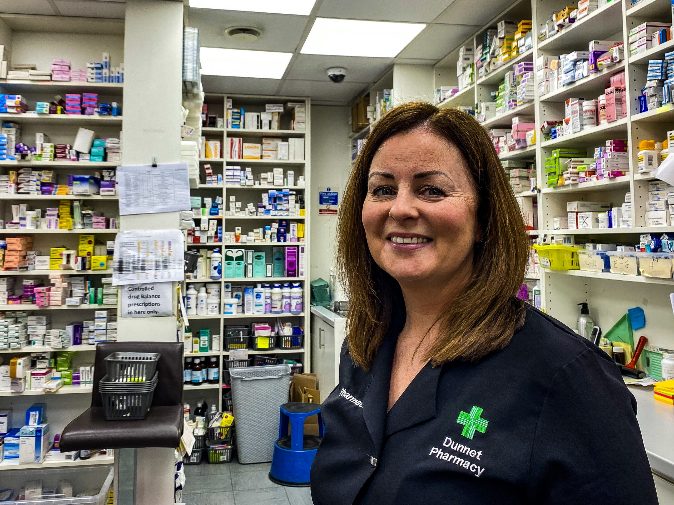 Maryann Dunnet of Dunnet Pharmacy in Glasgow, in the dispensing area of the pharmacy, with shelves of medicines behind her.