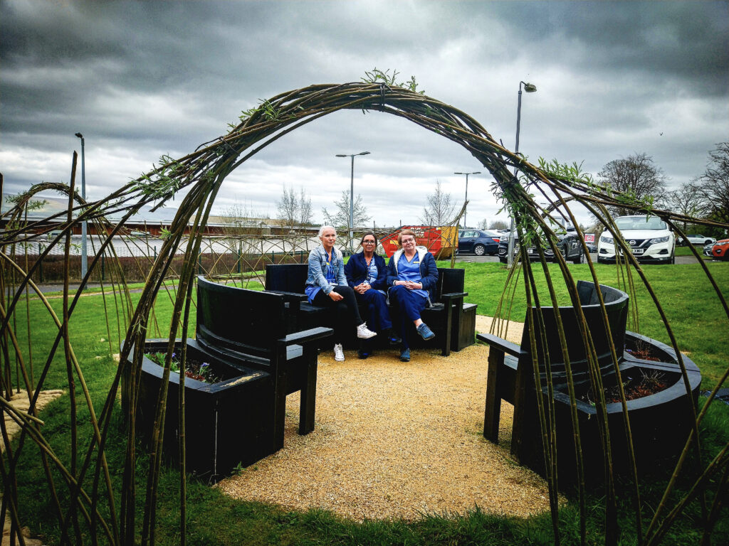 Three members of staff seated within the new Willow Arch surrounded by hospital grounds and under a cloudy sky