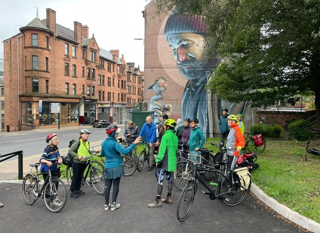 A group of cyclists stopped by the side of a Glasgow road next to one of its famous murals.